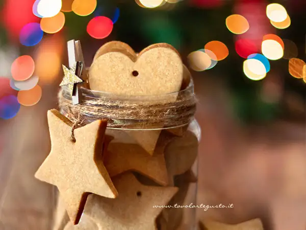 Biscotti alla cannella a forma di stella e cuore in un barattolo, decorati con luci natalizie sfocate.