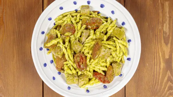 Piatto di trofie alla carlofortina con tonno, pomodorini secchi e pesto, servite su un piatto bianco con decorazioni blu.