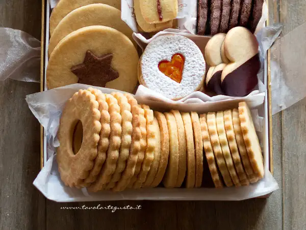 Scatola piena di deliziosi dolci senza glutine: biscotti rotondi con stella al cioccolato, a forma di cuore con zucchero a velo, ricoperti di cioccolato e impilati in file ordinate.